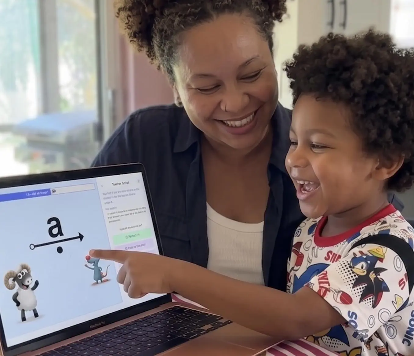 A smiling mother supporting her young son as he points to a laptop screen completing an interactive phonics reading lesson.