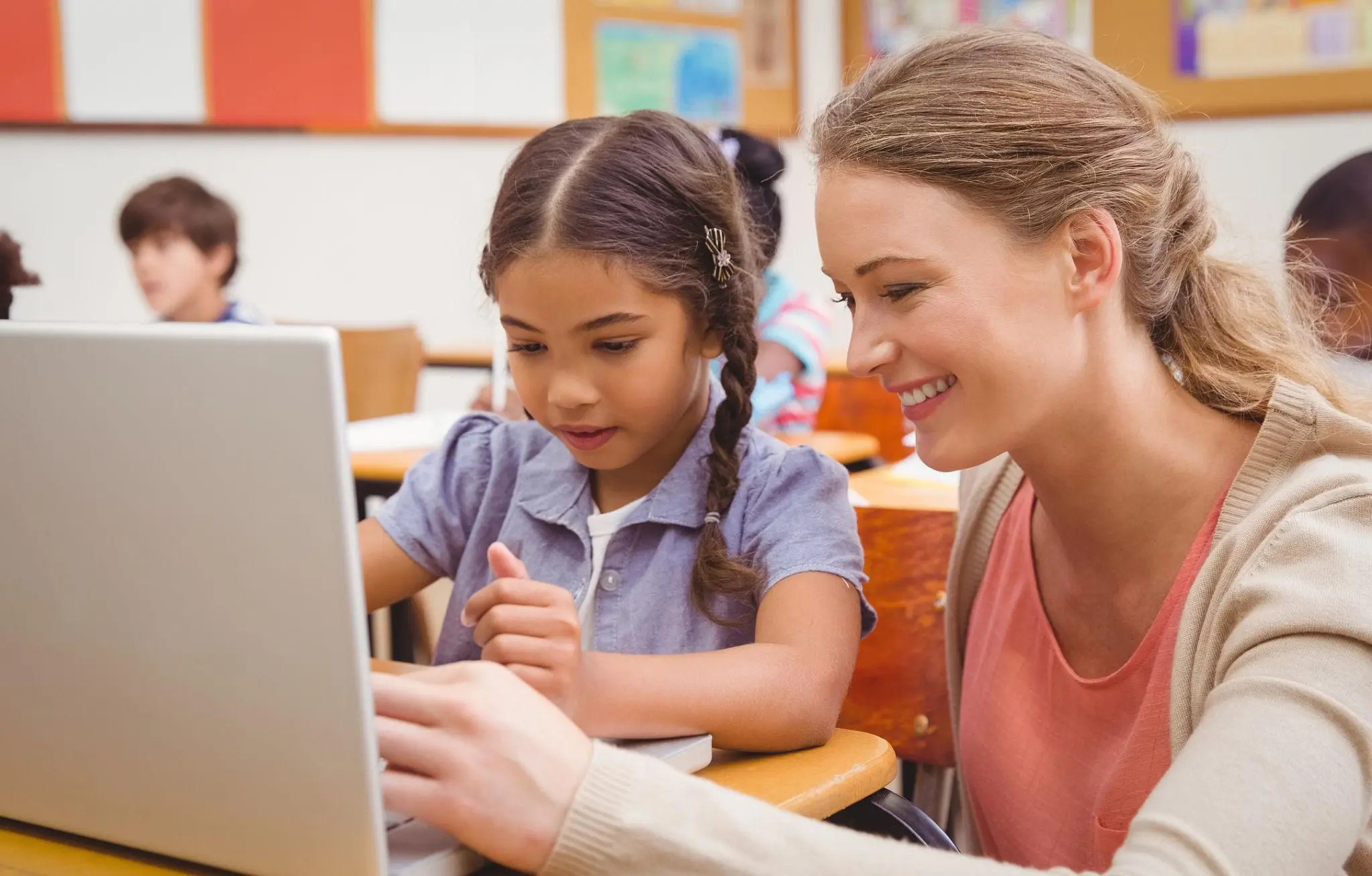A teacher smiling and sitting next to a young female student while helping her navigate a laptop lesson in a classroom.